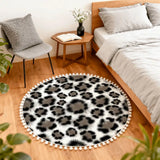 Gray leopard print round rug with pom poms beside a bed and small wooden side table